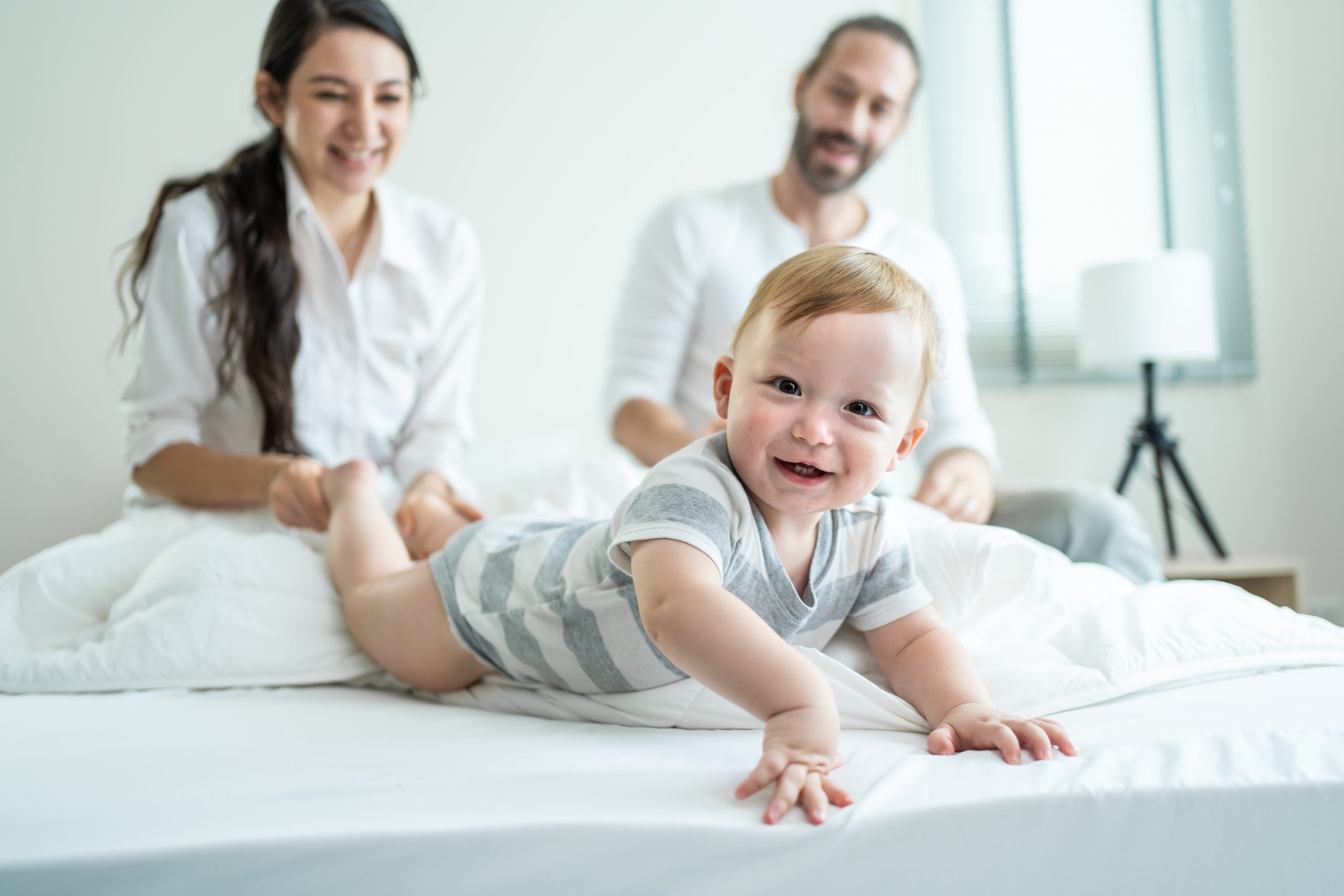 Portrait of Caucasian happy family smiling, look at camera in bedroom. Young attractive couple parents, father and mother sit on bed with little baby boy child enjoy morning wake up activity at home.