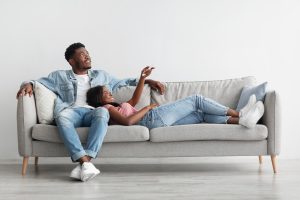 Cheerful Black Lady Holding Remote Controller, Opening Air Conditioner At Home While Relaxing On Comfortable Couch In Living Room, Happy Woman Adjusting Temperature Mode Lying On Guy's Lap, Free Space